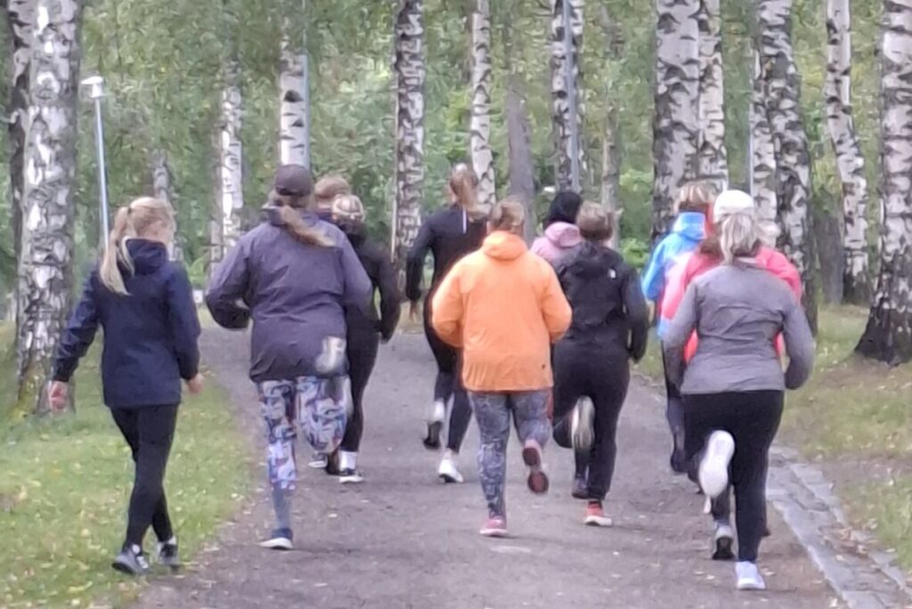 Kuvassa hiekkatiellä koivujen ympärillä juoksijoita kuvattuna takaapäin, valmentaja vasemmalla ottaa aikaa.